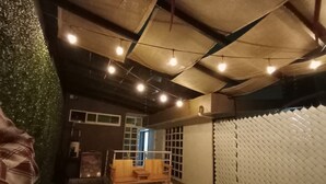 Quarto com carpete estampado, um banco de madeira, parede com padrão quadriculado e teto com vigas aparentes e luzes de cordão.
