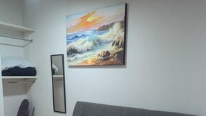 Quarto com sofá cinza, pintura de paisagem marítima, espelho e prateleira com roupas dobradas.
