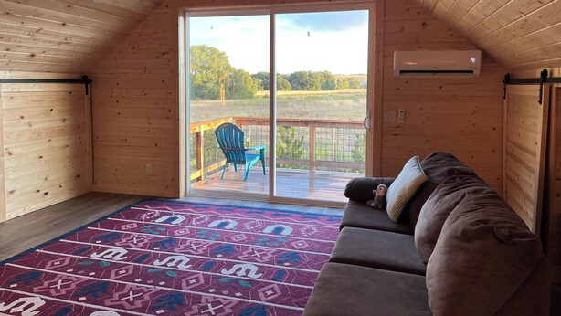 Ein gemütliches Blockhäuschen-Interieur mit Holzbalkendecke, einer Glastür zum Balkon und einem Teppich mit Muster sowie einem Sofa mit Kissen.