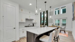 Une cuisine moderne avec des meubles blancs, un grand îlot central, des luminaires suspendus et un coin repas comprenant des chaises et une table.