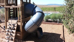 Un parque infantil con tobogán y zona de escalada, rodeado de un jardín ajardinado y vistas a montañas lejanas.