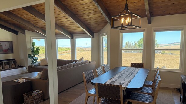 Amplia sala de estar con techo de madera, ventanas grandes que ofrecen vistas al campo, una mesa de comedor con sillas y un rincón de descanso acogedor con sofá y cojines.