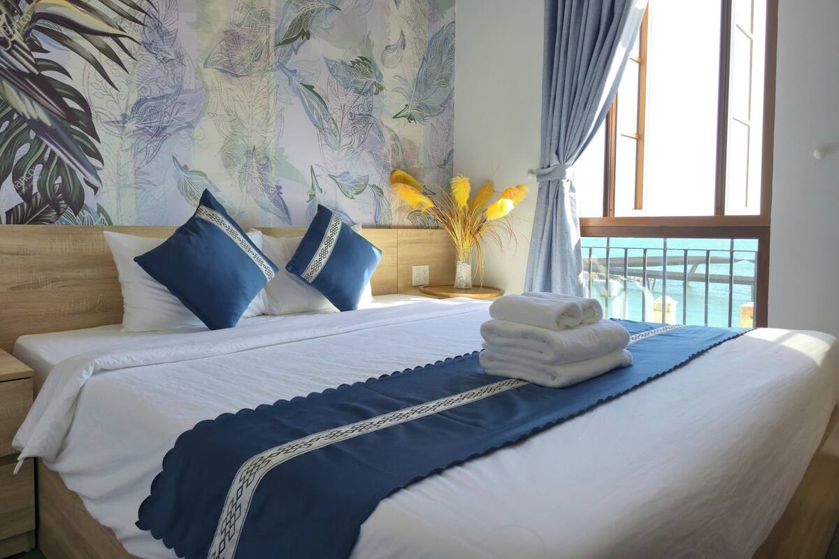 A hotel room with a large bed, a view of the sea through the window, and a tropical-themed wall mural.