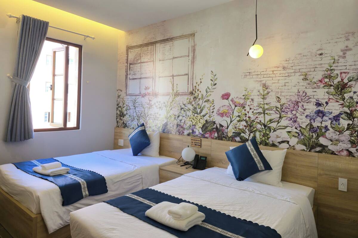 A hotel room with two beds, a floral mural, a window with curtains, and a bedside phone.