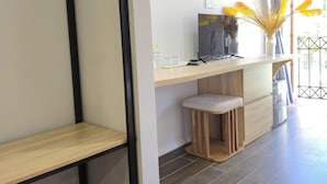 A modern living room with a wooden floor, a black metal shelf, a white wall with a framed picture, a small wooden stool, and a window with curtains. - Golden Sunset Hotel (Phu Quoc)