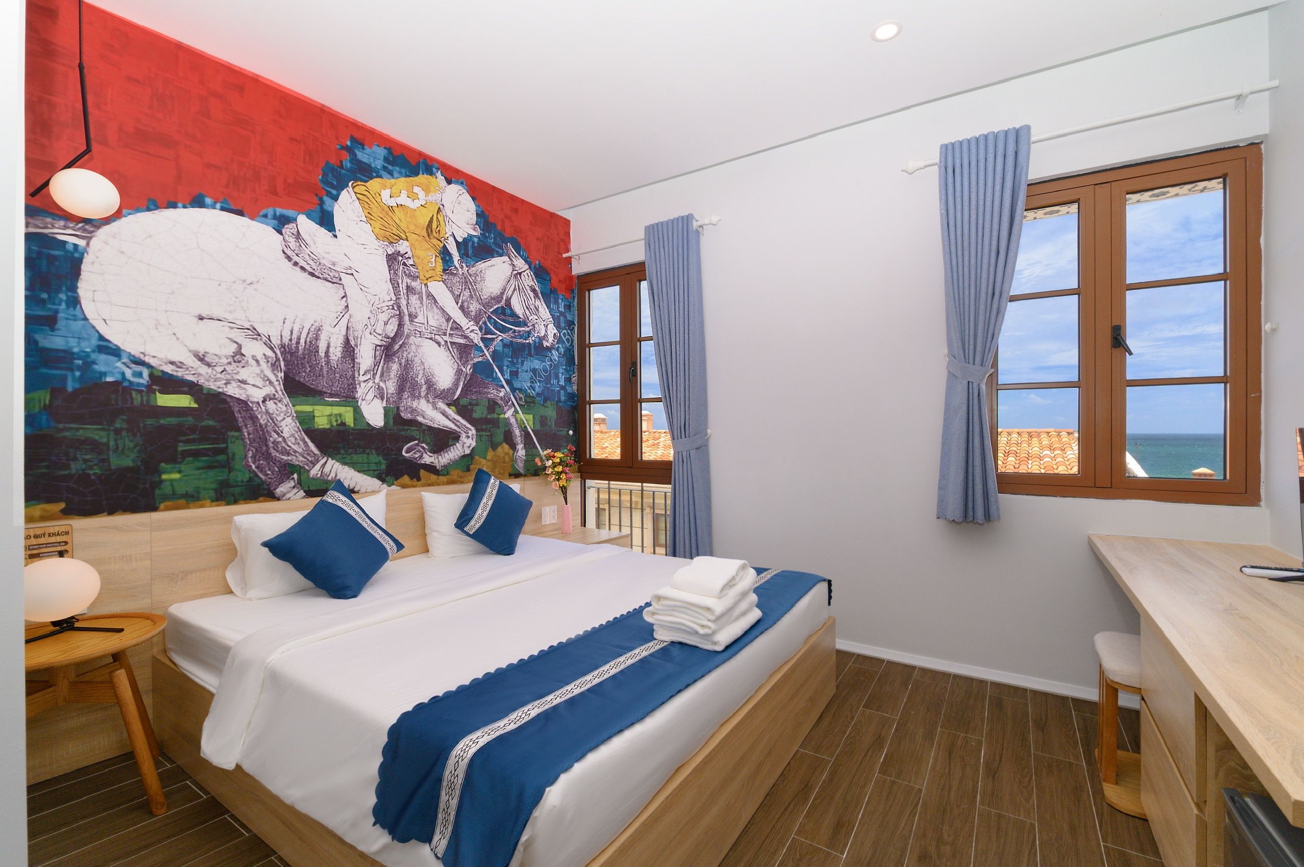 A hotel room with a bed, a desk, a window with curtains, and a mural on the wall.
