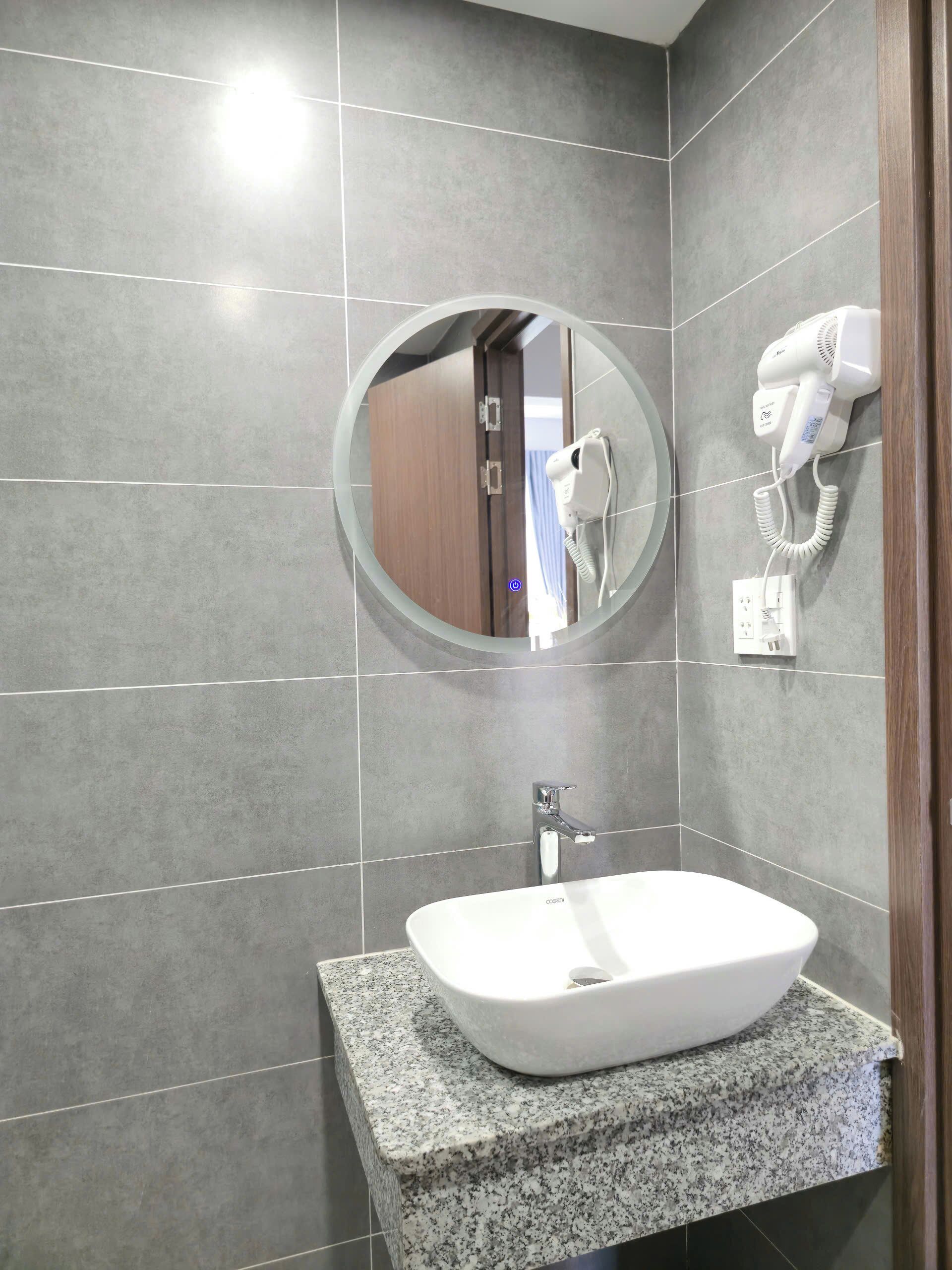 Un baño con encimera de granito, un lavabo blanco, un espejo redondo y un secador de pelo fijado en la pared.