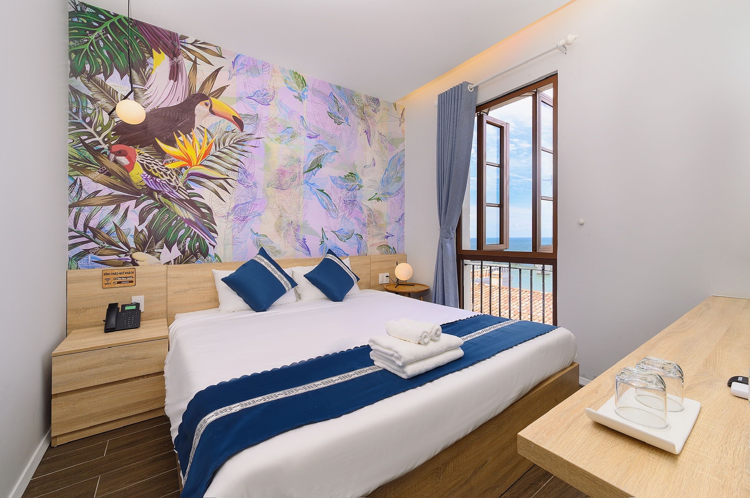 Een hotelkamer met een bed, een nachtkastje met telefoon, een raam met uitzicht op zee en een muur met een tropisch schilderij.