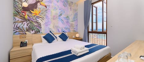A hotel room with a bed, a nightstand with a phone, a window with a view of the sea, and a wall with a tropical mural.