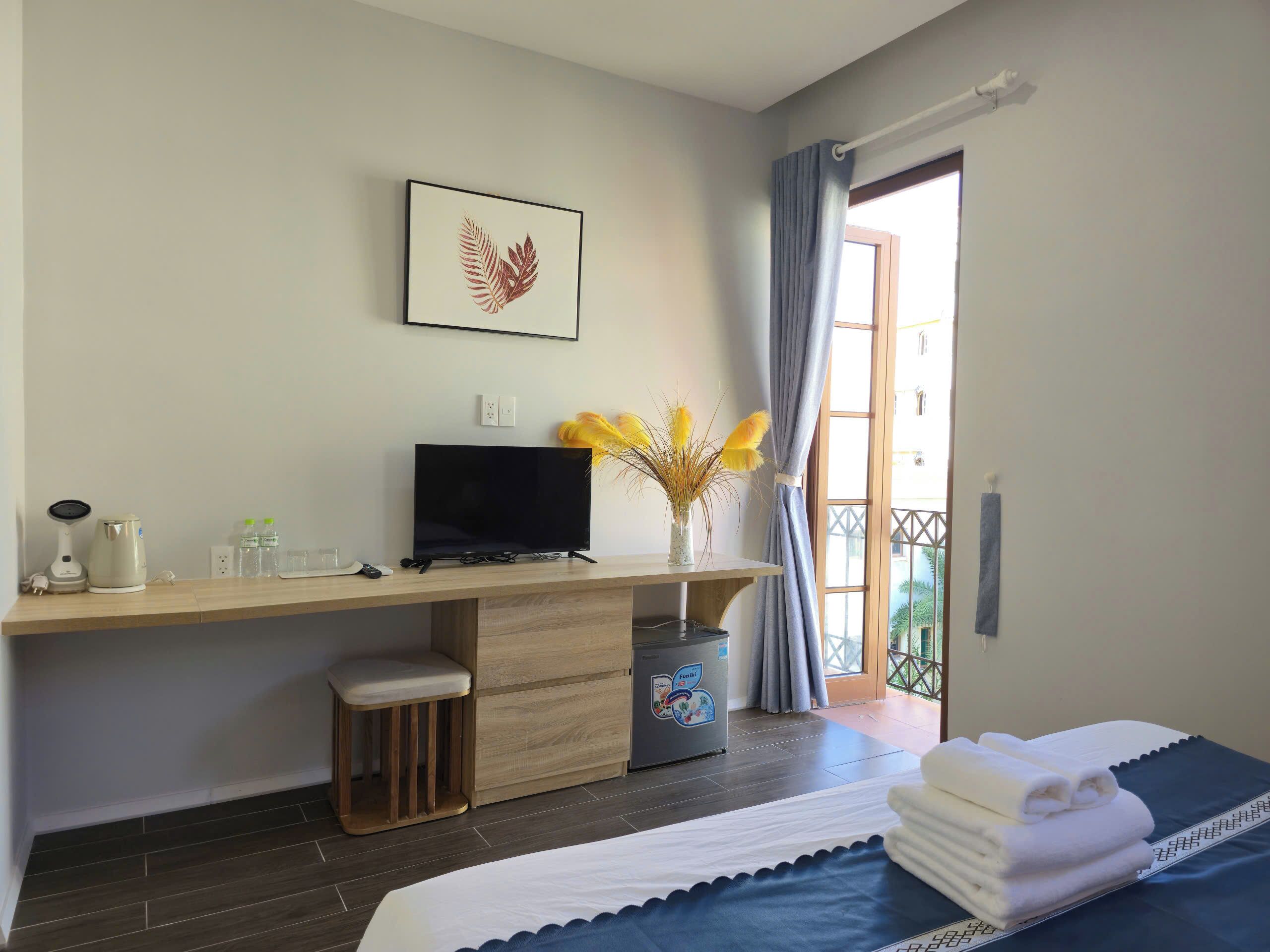 Habitación de hotel con cama, escritorio con televisor, taburete pequeño, armario y ventana con cortinas.