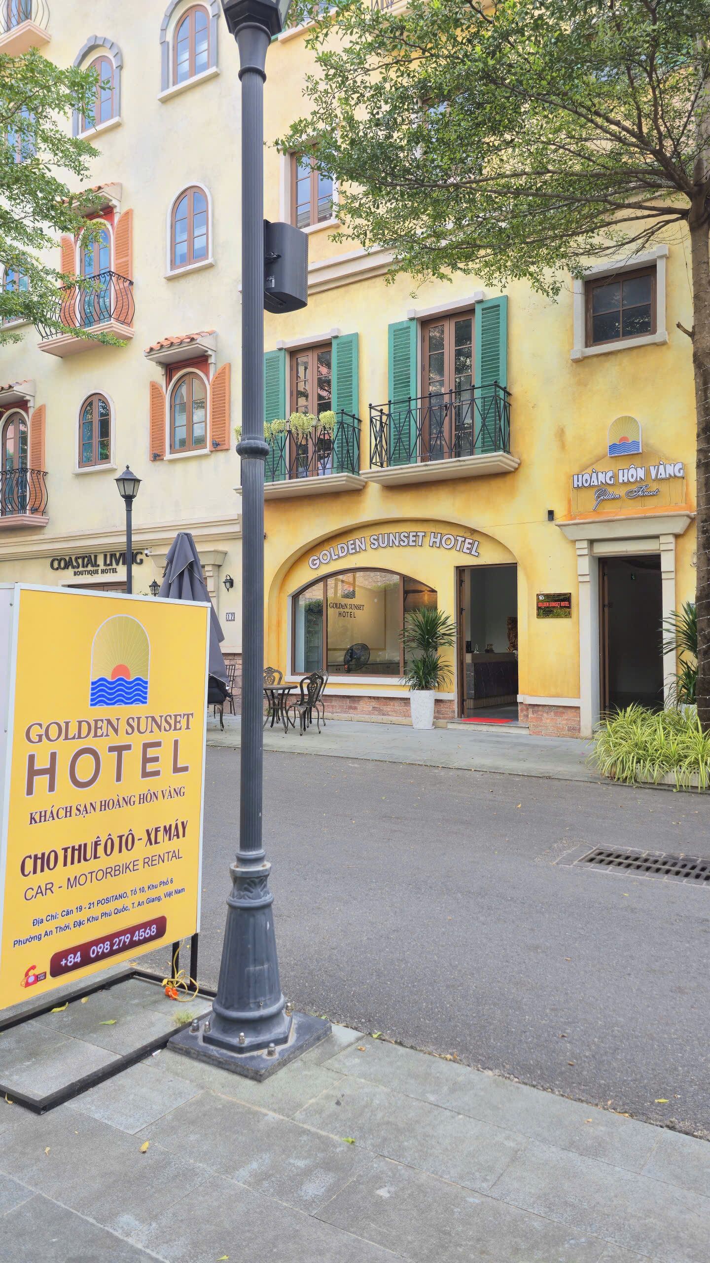 A hotel with a yellow facade, green shutters, and a signboard advertising car and motorbike rental services.