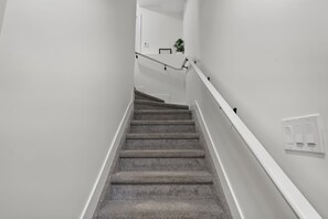 En trappe med tæppe beklædte trin og hvide gelændere, der fører op til en platform med en vægmonteret lyskontakt og en lille potteplante.
