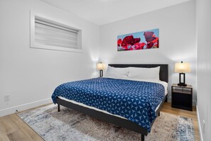 A bedroom with a bed, two bedside tables with lamps, a window with blinds, and a wall-mounted painting.