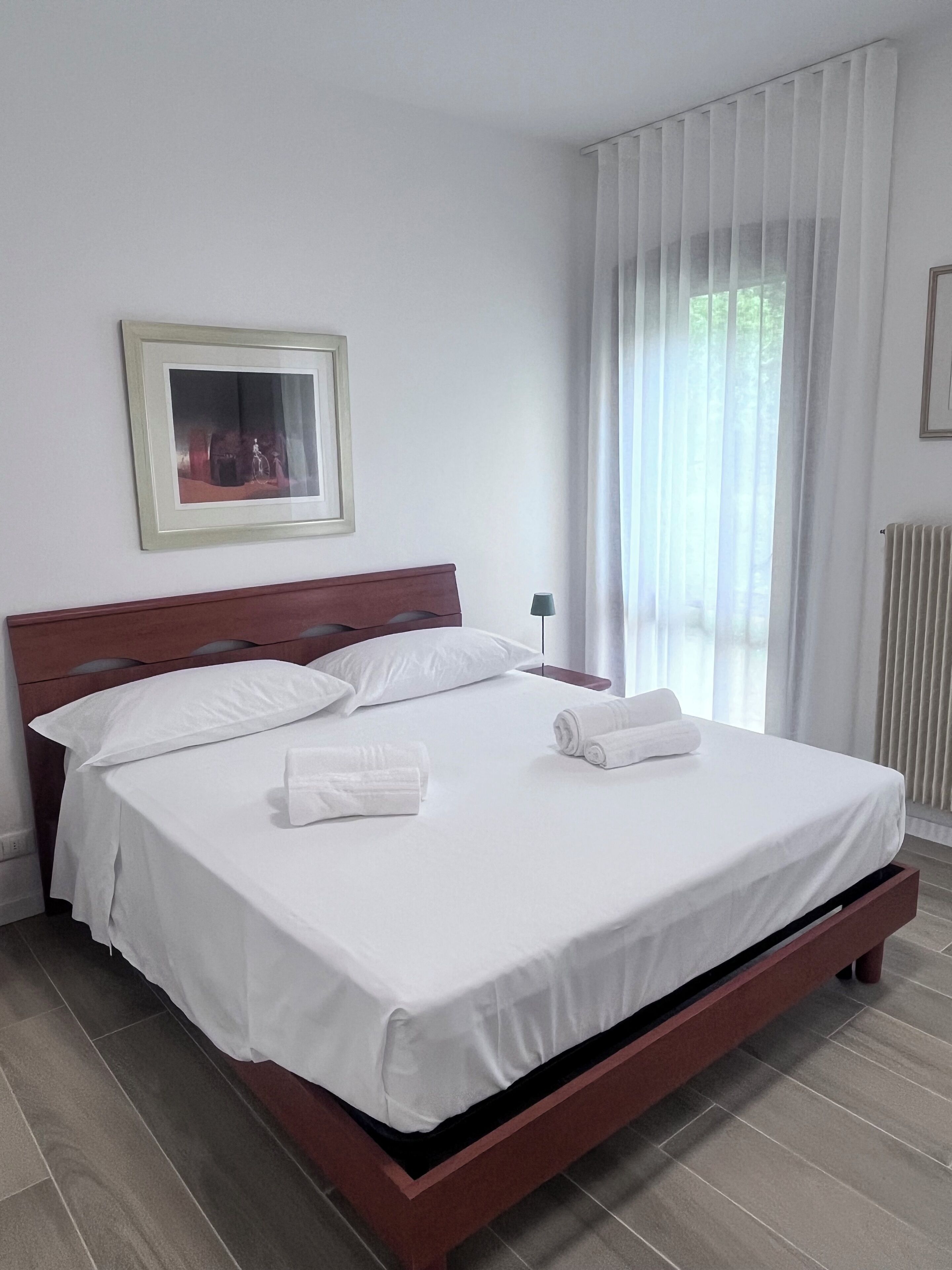 Ein ordentlich gemachtes Bett mit weißen Leinentüchern und zwei aufgerollten Handtüchern, ein Holzkopfbrett, ein gerahmtes Bild an der Wand und ein Fenster mit durchsichtigen Vorhängen.