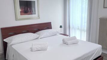 Ein ordentlich gemachtes Bett mit weißen Leinentüchern und zwei aufgerollten Handtüchern, ein Holzkopfbrett, ein gerahmtes Bild an der Wand und ein Fenster mit durchsichtigen Vorhängen.