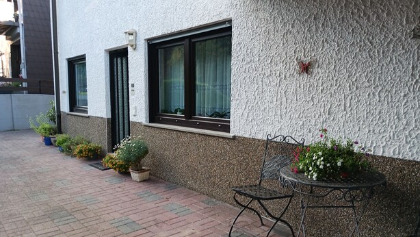 A building with a textured exterior, a small entrance, and a paved patio area with a metal table and chairs. - Vacation apartment in a quiet location (Friedrichsthal)