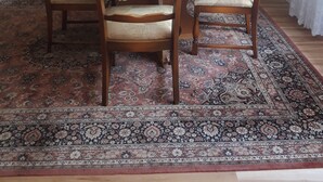A dining area with a round table, four wooden chairs, a patterned rug, a wooden cabinet with glass doors, and a vase with flowers on top. - Vacation apartment in a quiet location (Friedrichsthal)