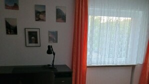 A room with a desk, chair, and a large window with curtains. There are pictures on the wall and a red rug on the floor. - Vacation apartment in a quiet location (Friedrichsthal)