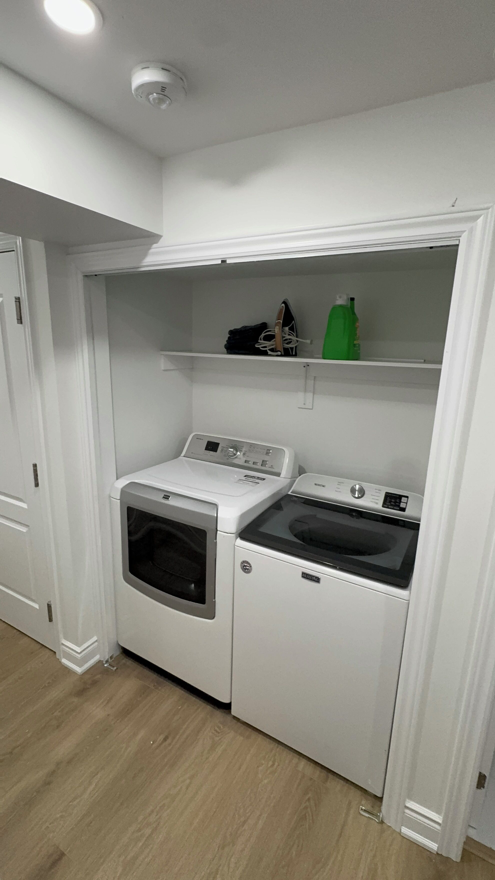 A laundry room with a front-loading washer and dryer, a shelf with a green bottle and a black item, and a white door.
