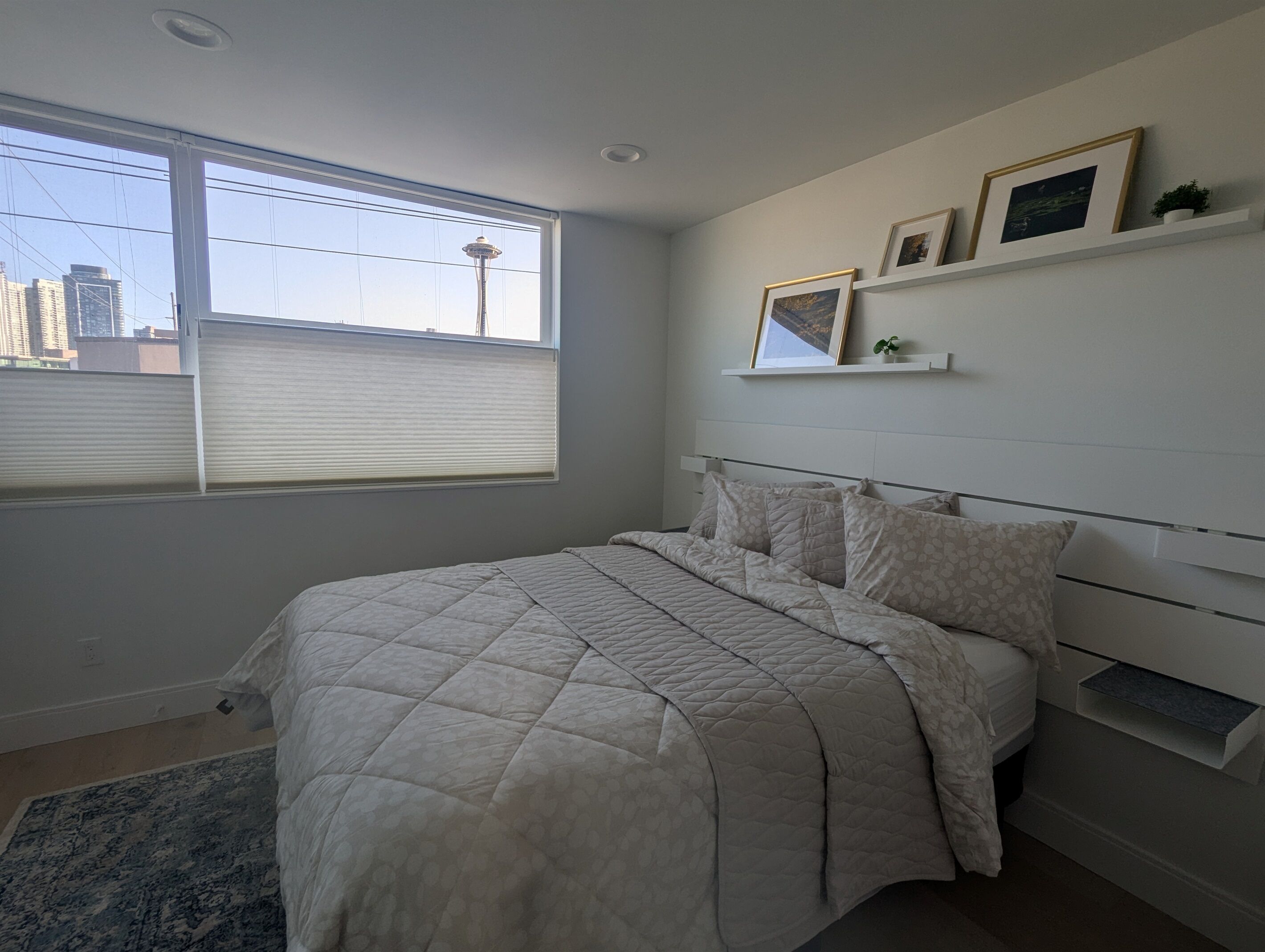 A bedroom with a large bed, a window with blinds, a city view, and a shelf with framed pictures.