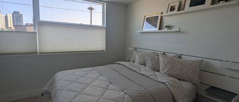 A bedroom with a large bed, a window with blinds, a city view, and a shelf with framed pictures.