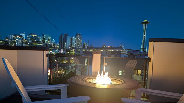 A rooftop terrace with a fire pit, two white Adirondack chairs, and a city skyline view including the Space Needle.