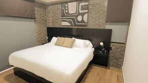 A modern bedroom with a brick accent wall, a large bed with white bedding, and a nightstand with a lamp.