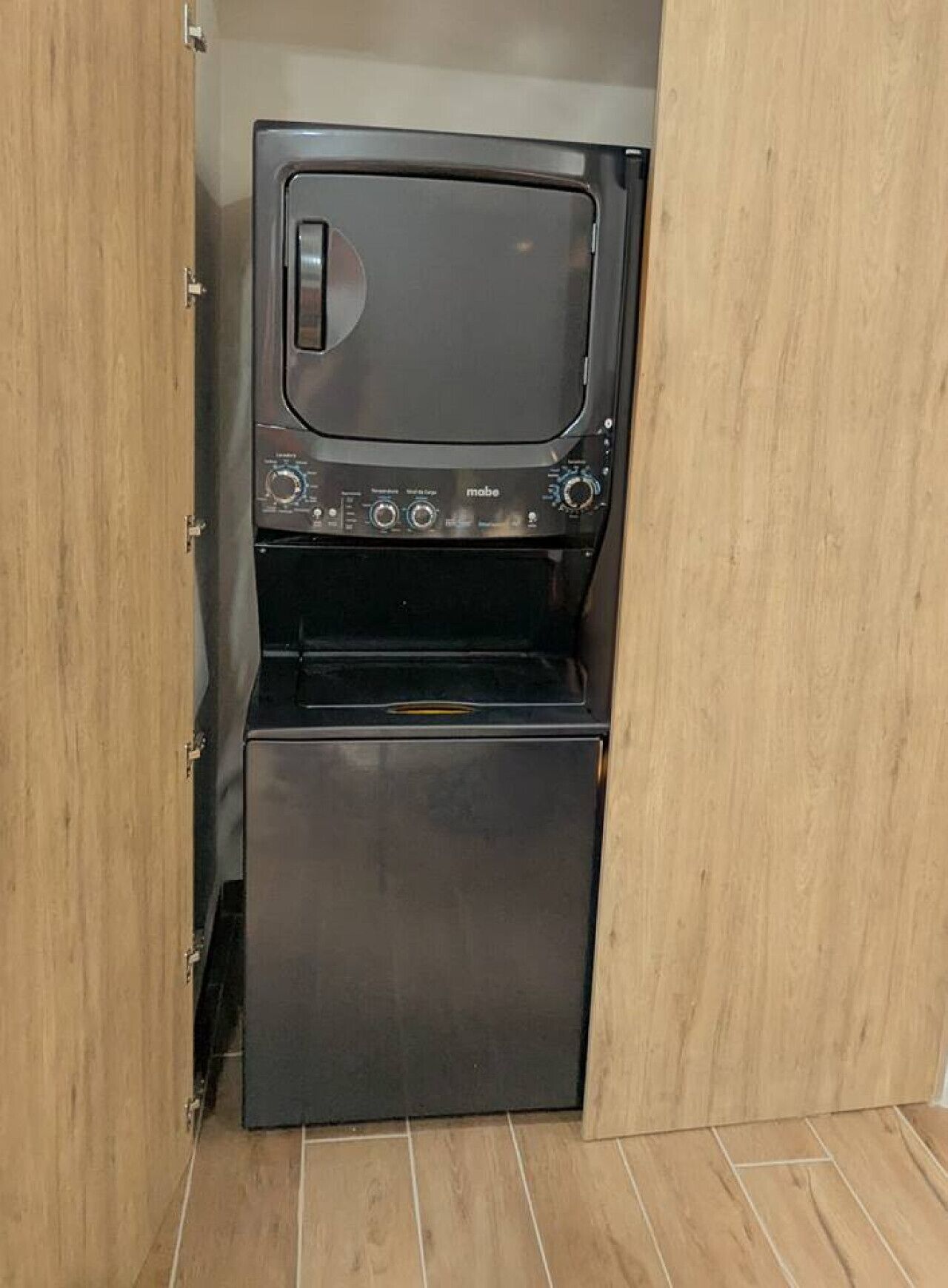 Una lavadora compacta de color oscuro con un panel de control, ubicada en una habitación con piso de madera y una puerta entreabierta.