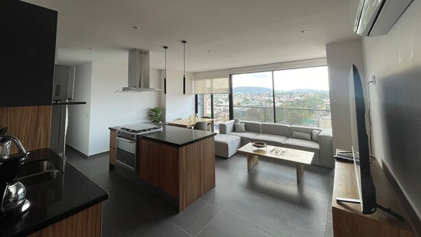 A modern kitchen with a central island, a dining area with a sofa and coffee table, and a large window offering a city view.