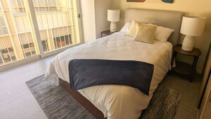 A hotel room with a large bed, two bedside tables with lamps, a large window with a view of buildings, and a modern abstract painting on the wall. - Beautiful Apartment in the Heart of Zapopan 306A (Zapopan)