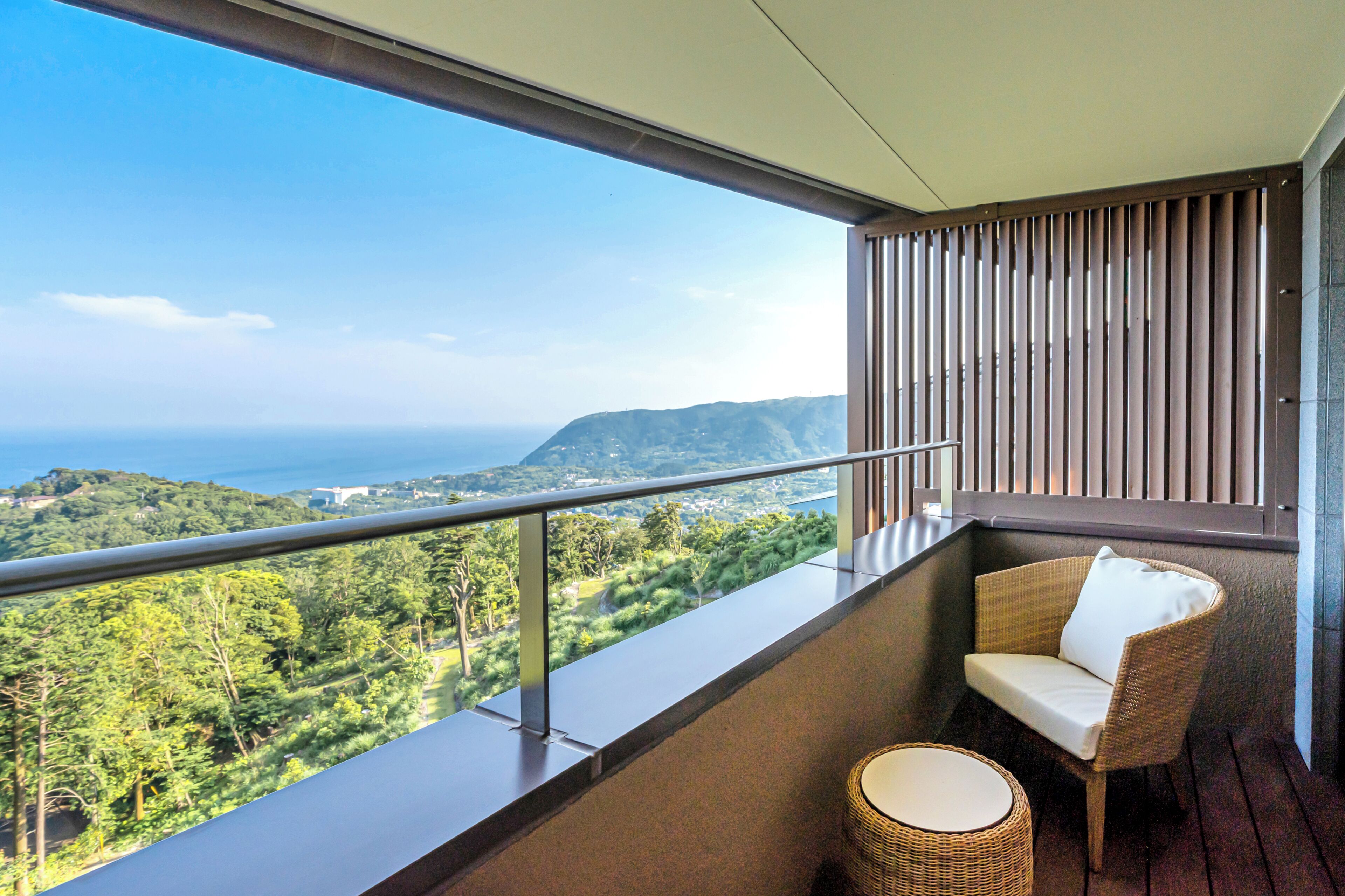 A balcony with a wicker chair and a small table, overlooking a lush landscape and the sea.