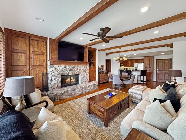 A spacious living room with a stone fireplace, wooden beams, a ceiling fan, and a dining area with a kitchen in the background.
