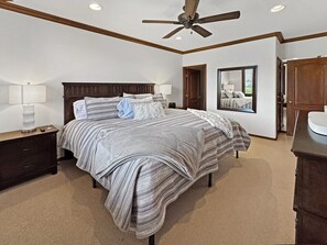A bedroom with a large bed, wooden headboard, bedside tables with lamps, a ceiling fan, and a view through a window.