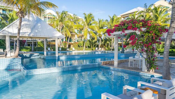 Ein Swimmingpool mit Liegestühlen und einer überdachten Liegefläche, umgeben von Palmen und einem Gebäude mit weißer Fassade.