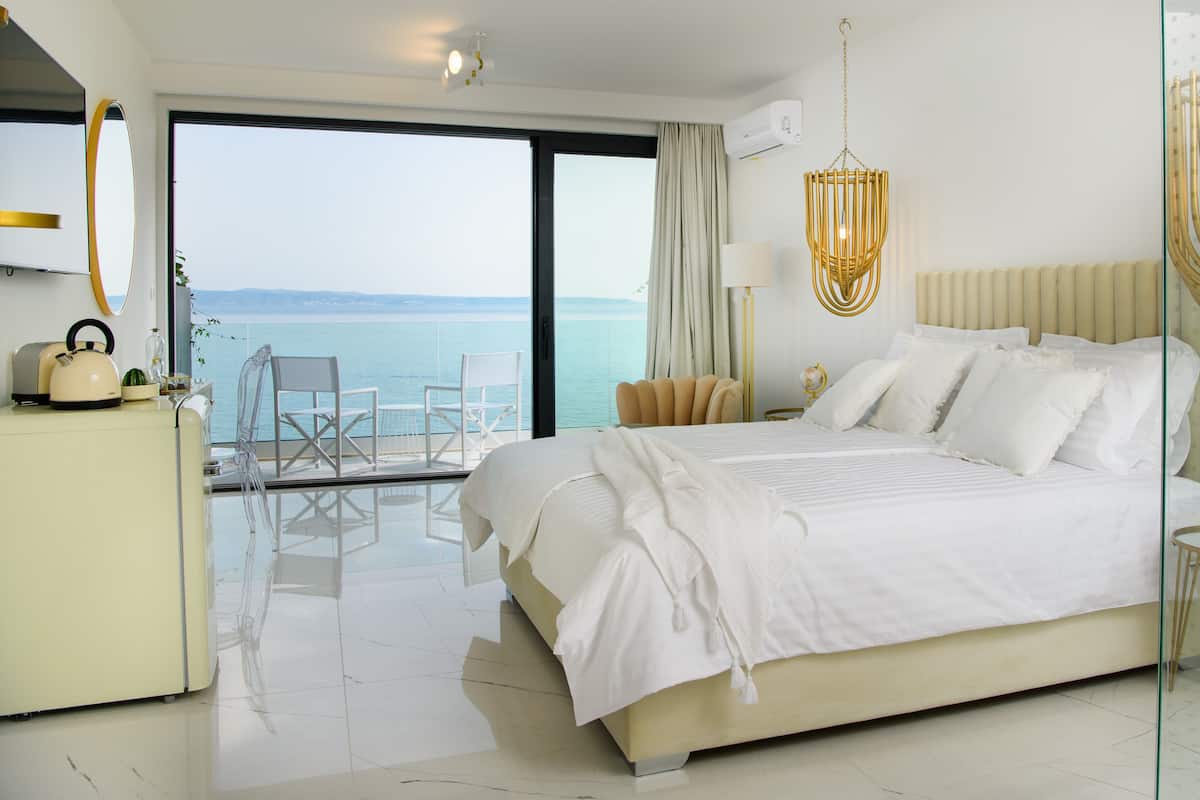 Una habitación de hotel moderna con una cama grande, un balcón con vistas al mar, un área de descanso con una silla y una mesita con un hervidor de agua.