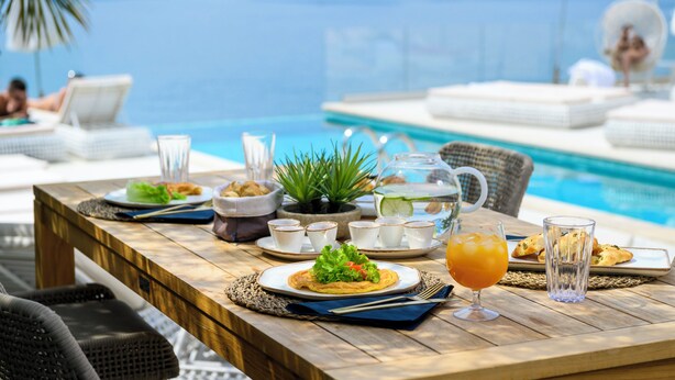 Una mesa de comedor con platos de comida, vasos y una jarrón de jugo junto a una piscina con vista al mar.