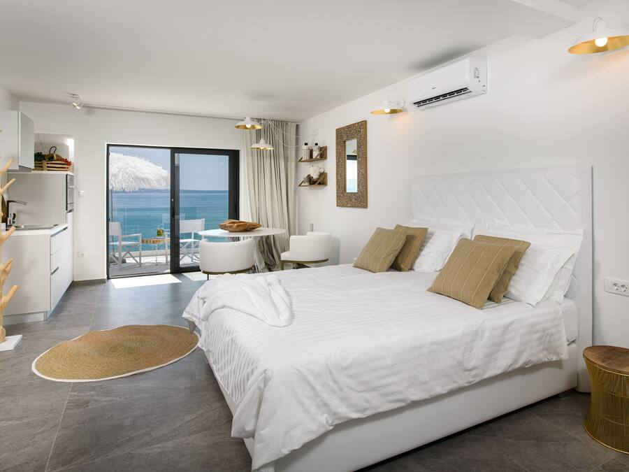 A modern bedroom with a large bed, a view of the sea, a balcony, and a kitchenette.