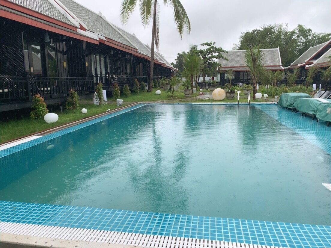 A swimming pool with a tiled edge, surrounded by a covered outdoor area with lounge chairs and a building with a sloped roof.