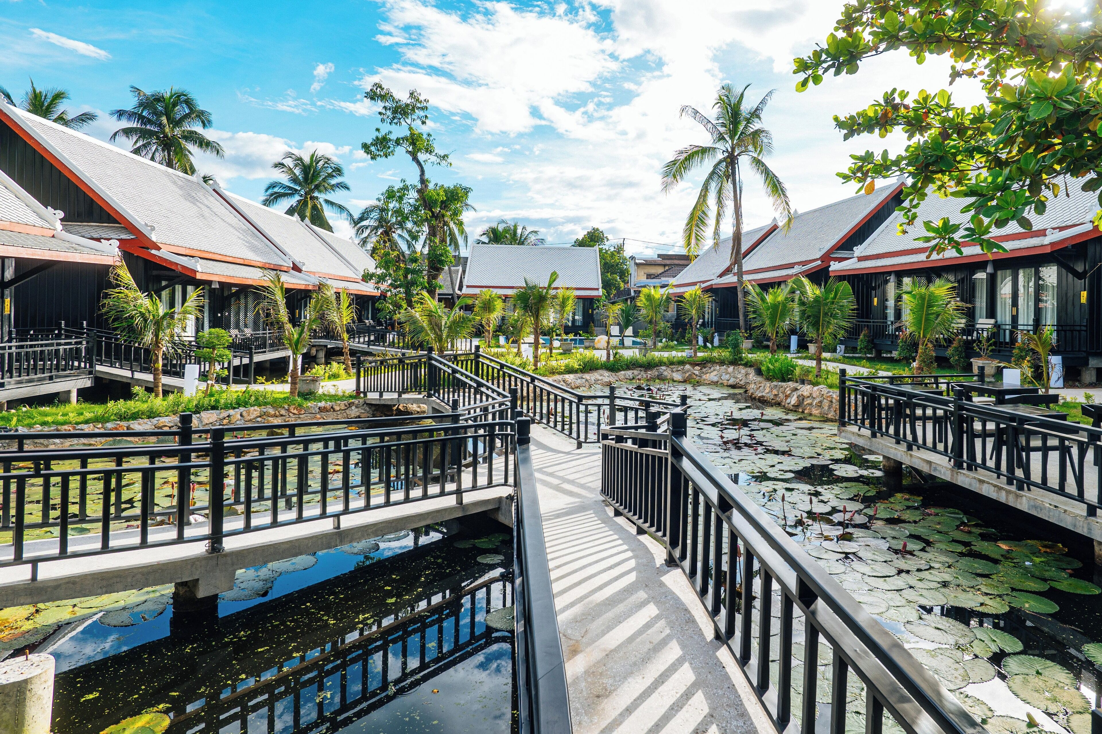 A resort with multiple buildings, a bridge over a pond with lily pads, and palm trees.