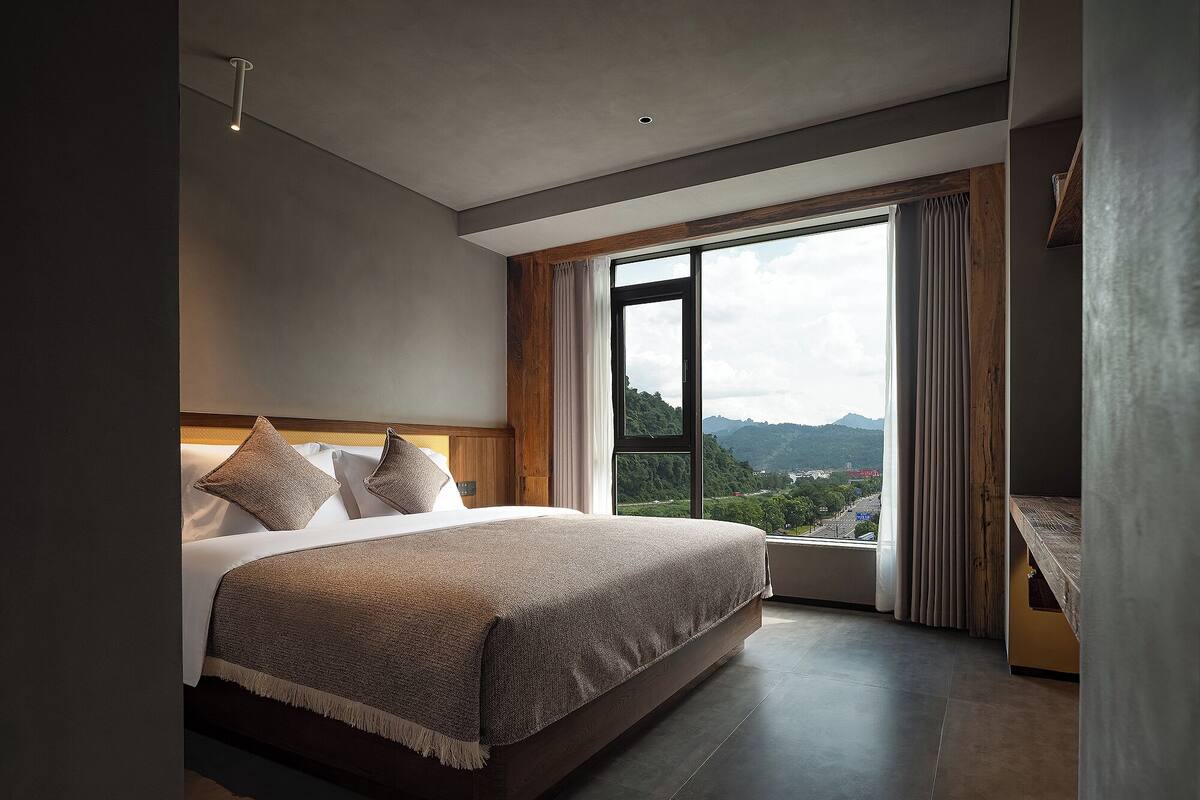 A modern bedroom with a large bed, a view of the outdoors, and a wooden headboard.