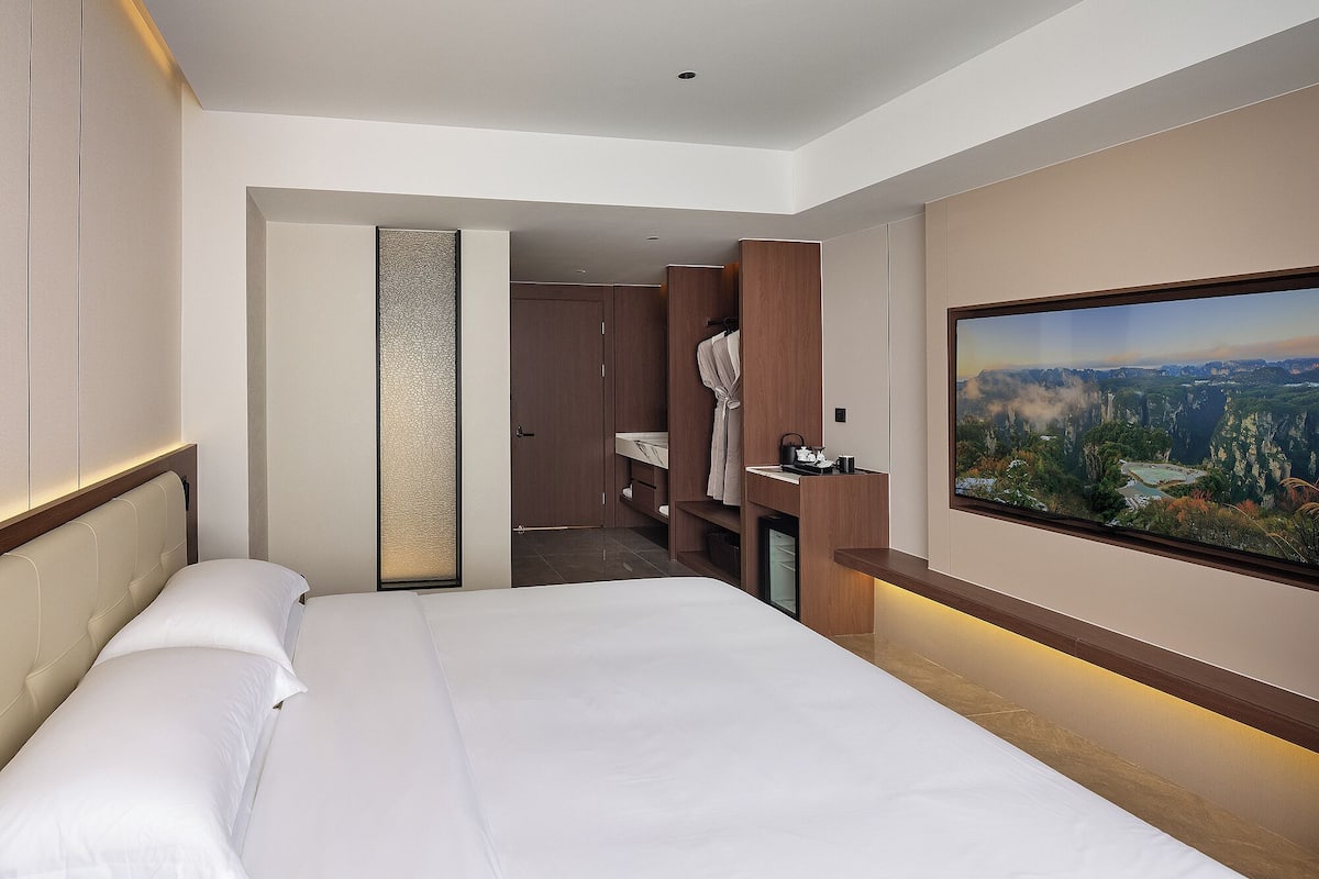 A hotel room with a large bed, a bathroom with a shower, a vanity, and a large window with a mountain view.