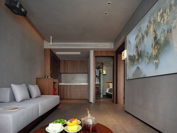 Una habitación de hotel moderna con un sofá gris, una mesa de comedor con fruta y té, una zona de cocina y un cuadro grande de un paisaje montañoso.