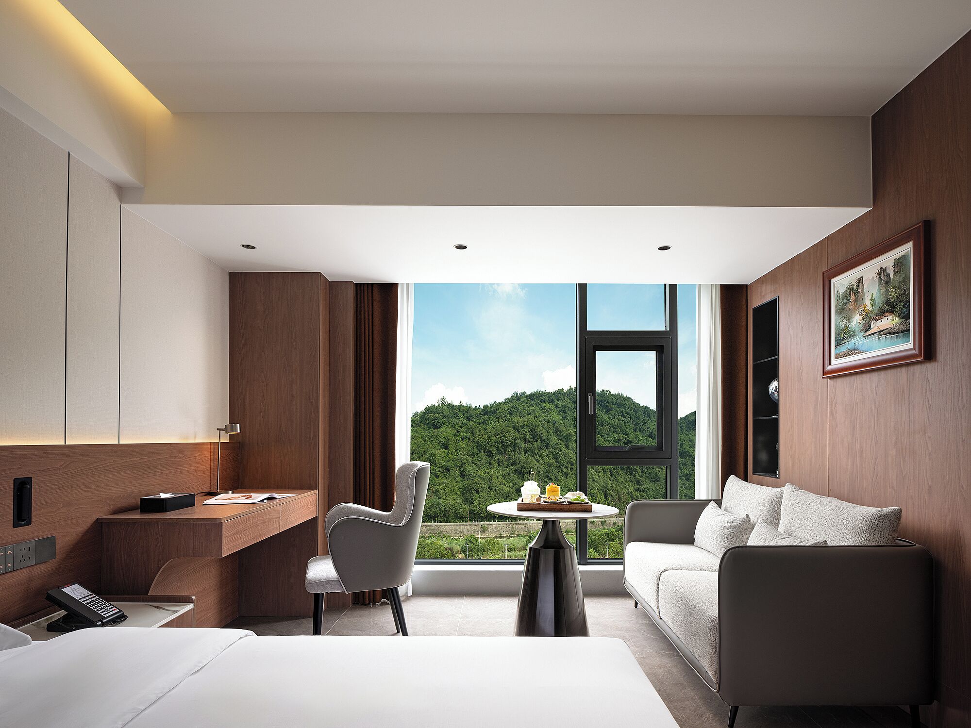 A modern hotel room with a large window, a desk, a chair, a sofa, and a small table.