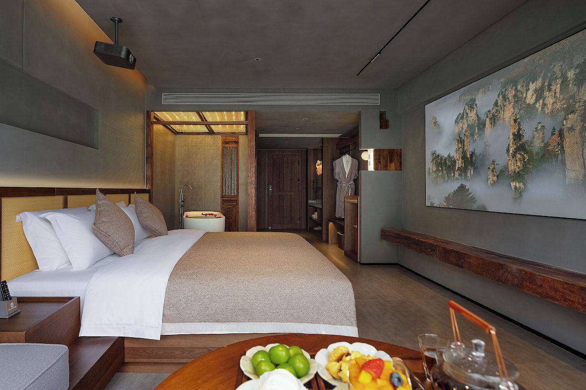 A modern hotel room with a large bed, a dining table with fruit, a robe, and a painting of a mountainous landscape.
