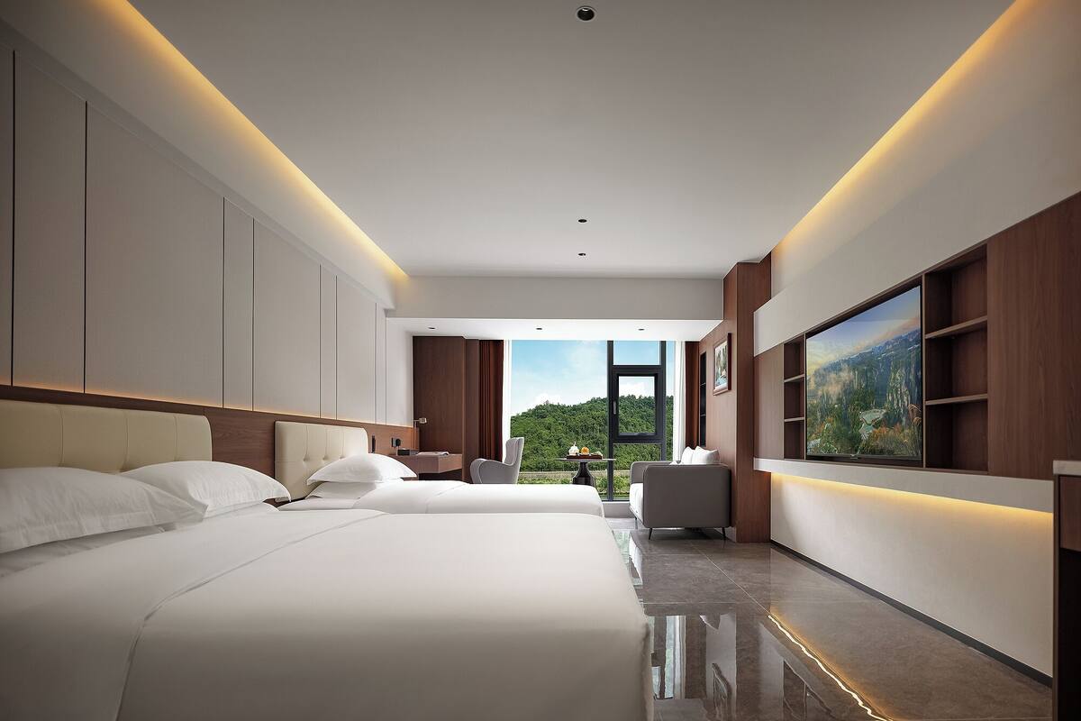 A modern hotel room with two beds, a desk, a chair, and a large TV on the wall.