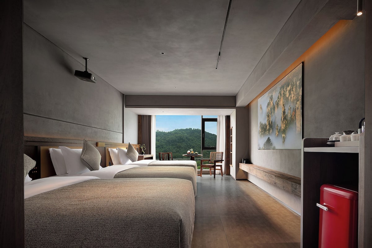 A modern hotel room with two beds, a dining area, and a view of the outdoors.