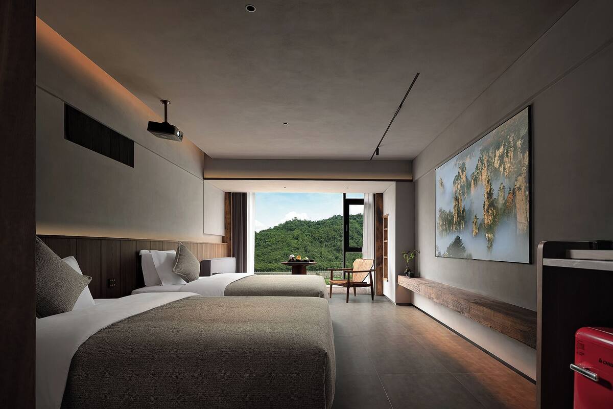 A modern hotel room with two beds, a large window with a mountain view, and a painting on the wall.