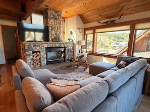 A cozy living room with a stone fireplace, a large grey sofa, and a view of a mountainous landscape through large windows.