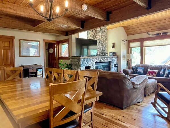 A spacious living room with a wooden dining table and chairs, a stone fireplace, and a comfortable sofa set. - Big Sky Resort - Ski in and out Arrowhead Chalet 1657 (Big Sky)