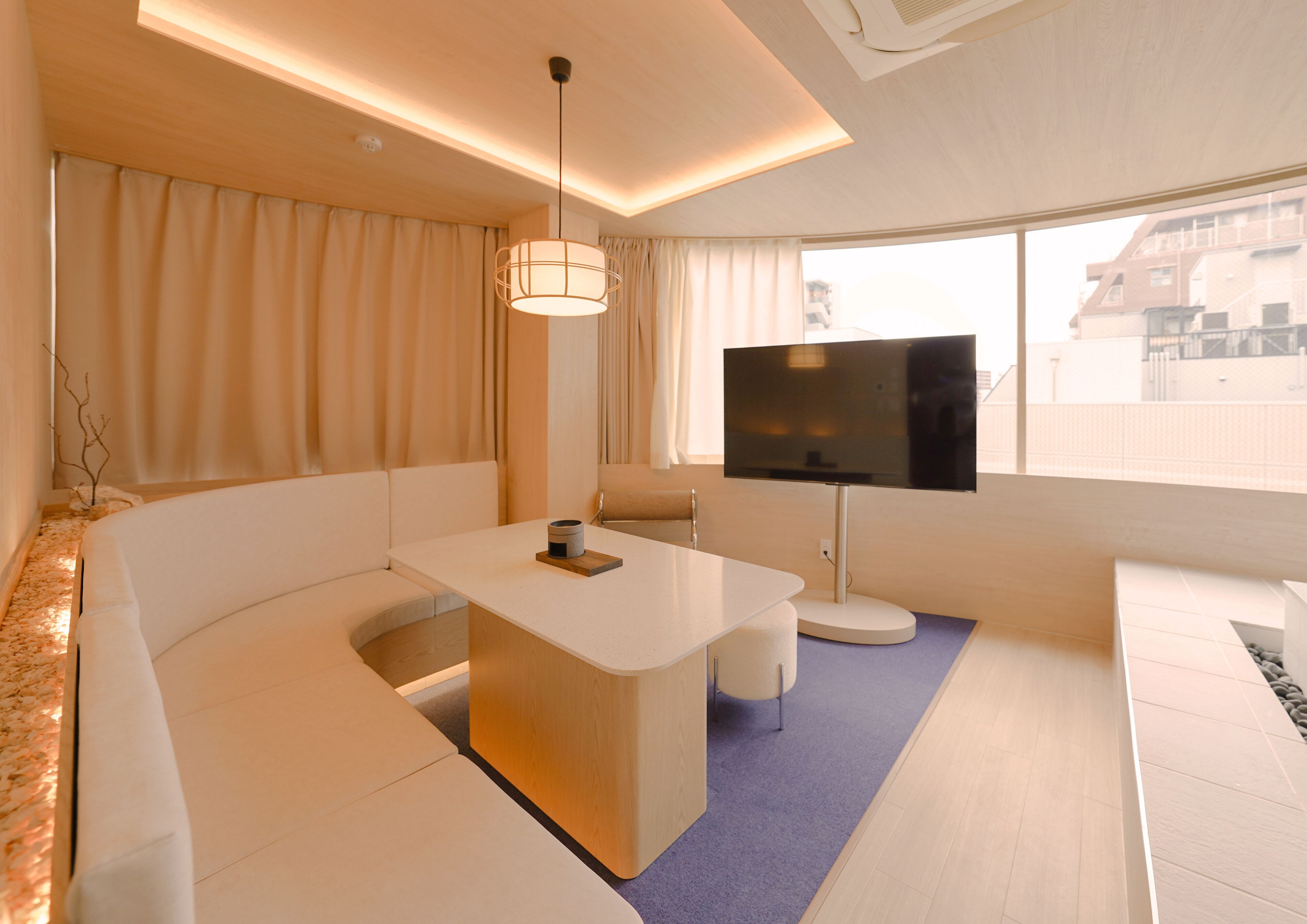 A modern hotel room with a sofa, a small table, a TV, and a window with a city view.